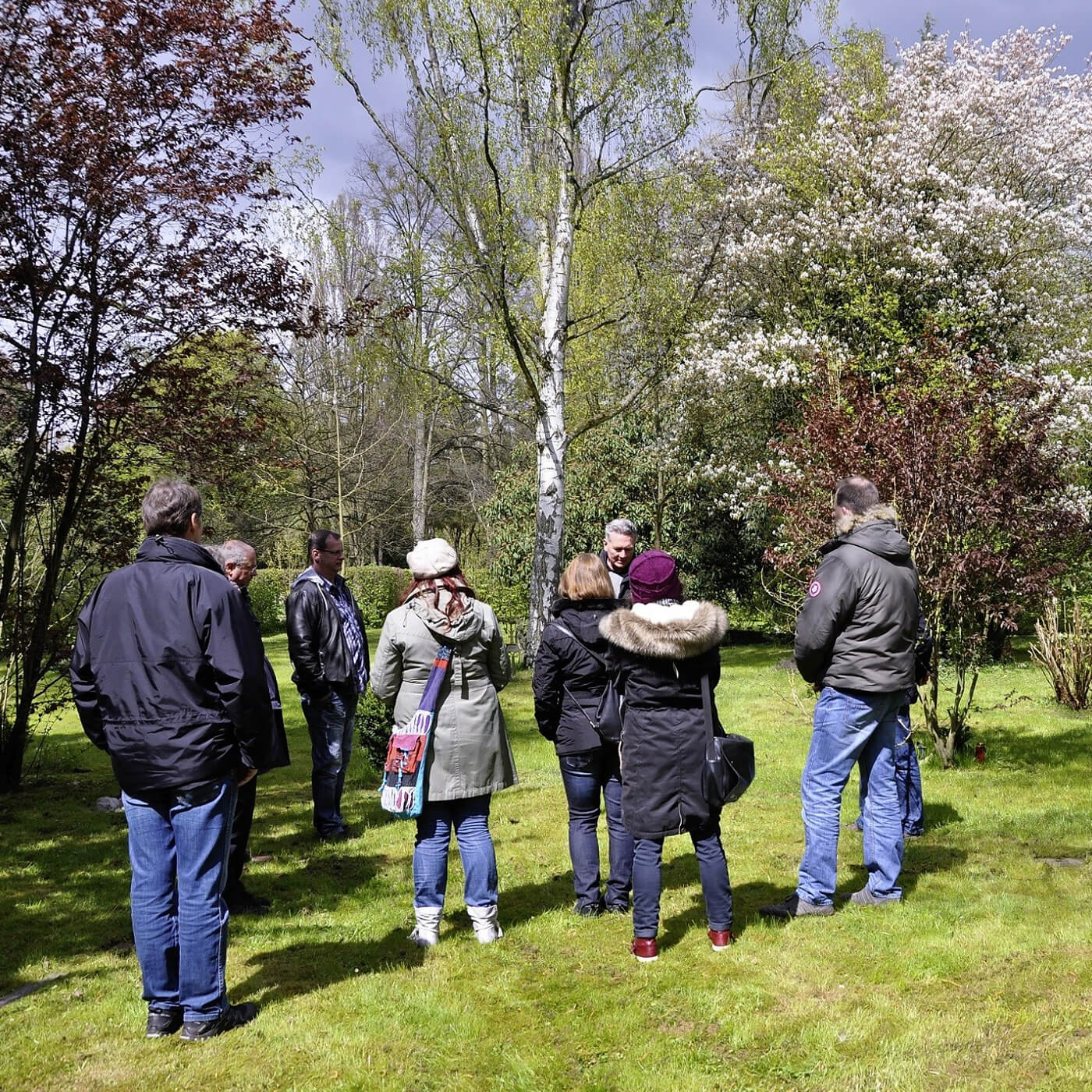 Gruppe von acht Personen steht auf einer Wiese umgeben von Bäumen