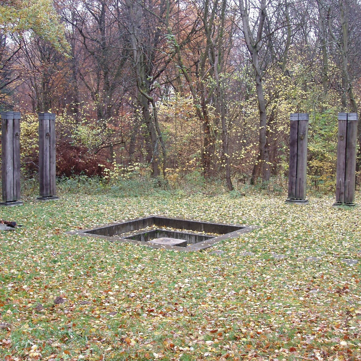 Vier hohe, rechteckige Holzsäulen stehen auf einer mit Herbstlaub bedeckten Wiese um eine rechteckige Vertiefung aus Beton