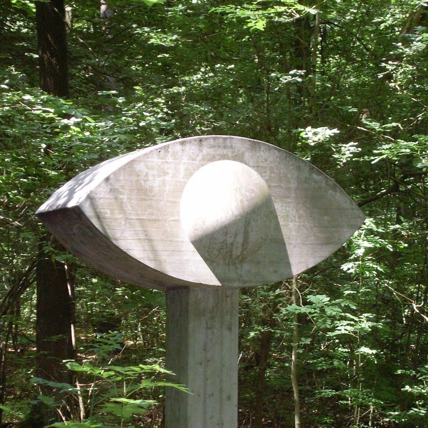 Betonskulptur in Form eines stilisierten Auges auf einem Pfeiler im Wald