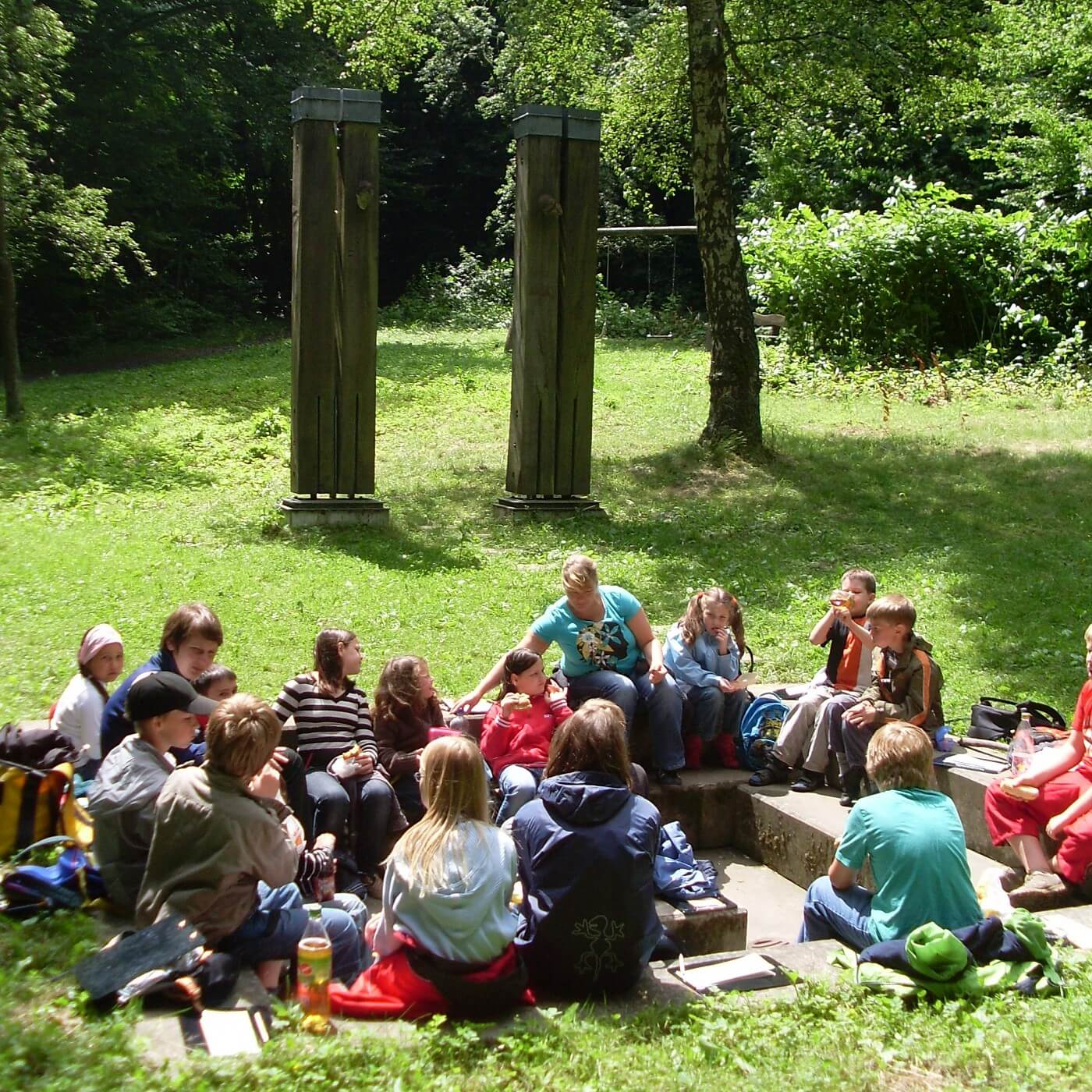Gruppe von Kindern sitzt im Halbkreis auf einer Wiese vor zwei hohen Holzskulpturen