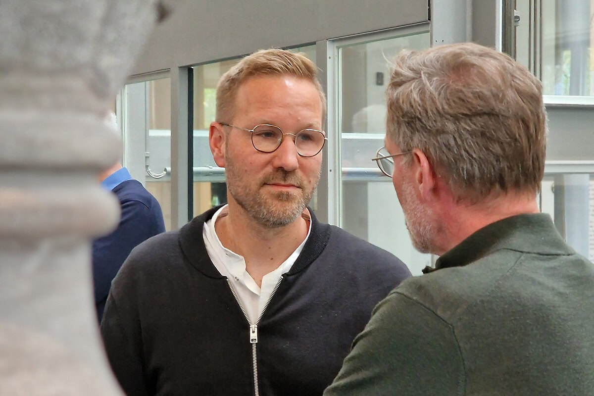 Zwei Männer im Gespräch in einem hellen Raum mit großen Fenstern, einer trägt eine Brille und einen grünen Pullover, der andere einen schwarzen Reißverschluss-Pullover über einem weißen Hemd