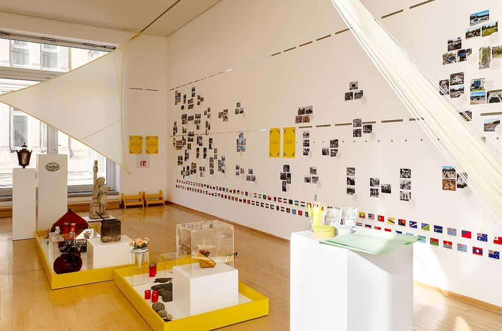 Ausstellungsraum mit einer Wand voller Fotos, Flaggen und Vitrinen mit Objekten
