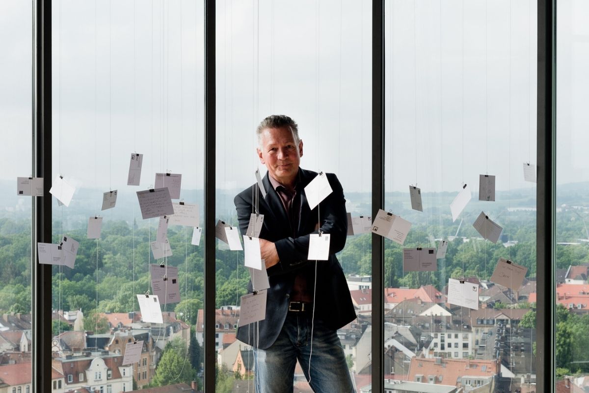 Museumspädagoge Gerold Eppler steht in der Rotunde des Museums für Sepulkralkultur. Um ihn herum sind an schnüren Postkarten aufgehängt. Sie scheinen zu schweben. Hinter ihm sieht man durch die Fenster die Stadt Kassel. Er verschränkt die Arme.