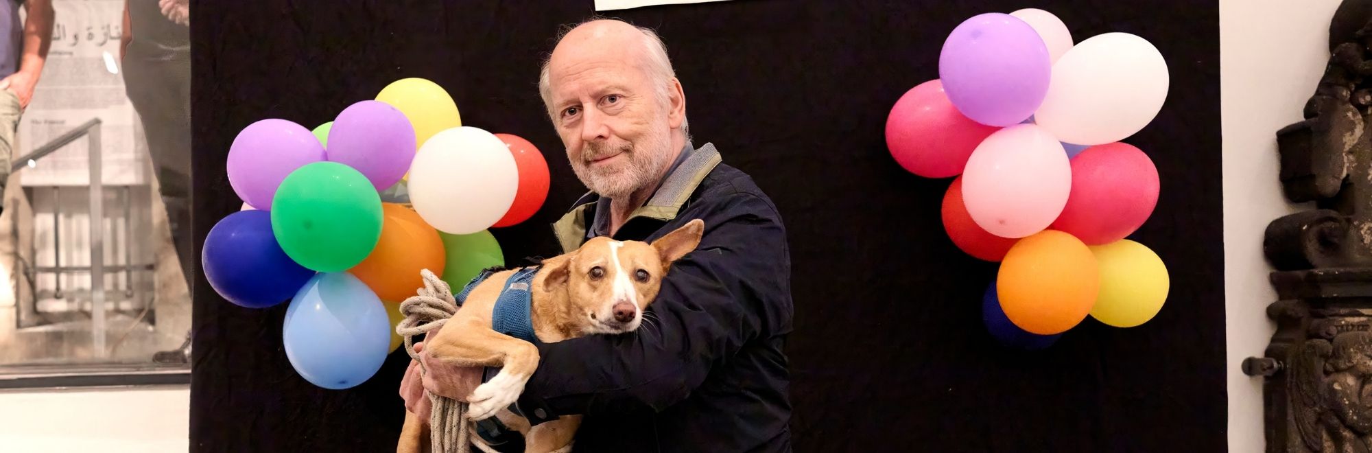 Ein älterer Mann hält einen Hund im Arm. Im Hintergrund bunte Luftballons.