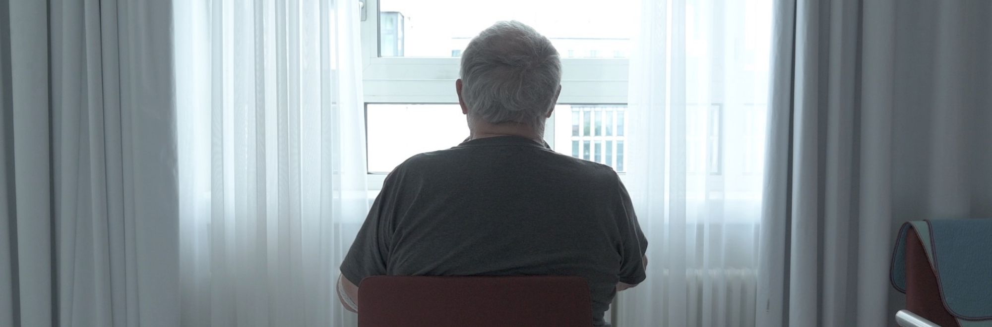 Auf einer Palliativstation. Ein alter Mann sitzt mit dem Rücken zum Betrachter auf einem Stuhl und schaut aus dem Fenster.